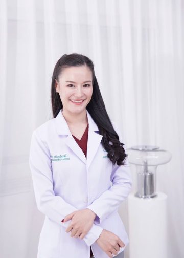 พลอย ศิริอร ยิ้ม สวมเสื้อกาวน์สีขาวทับชุดสีแดงเลือดหมู ยืนหน้าผ้าม่านสีขาว